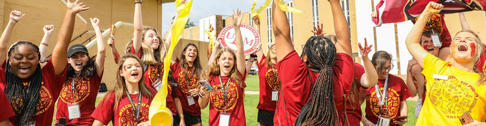 photo of about a dozen ɫAV students wearing matching marron and gold shirts cheer with their arms raised. With streamers and flags flying, the students exude high energy on a grassy lawn with yellow brick buildings in the background.